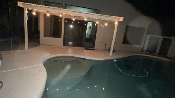 Una piscina con un porche cubierto, luces colgantes y una casa al fondo.