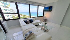 A modern living room with a white sofa, a glass coffee table, and a view of the ocean. - Modern 3 Bedroom with Ocean Views - 1104 (Miami Beach)