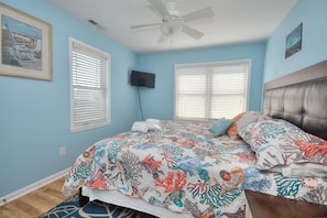 A bedroom with a bed, a ceiling fan, two windows with blinds, a mounted TV, and a framed picture on the wall.