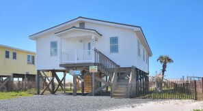 A white two-story house with a balcony, elevated on stilts, and a wooden fence in the background.