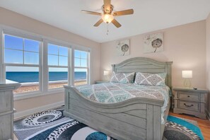 A bedroom with a bed, a nightstand, a lamp, a ceiling fan, and a view of the ocean through large windows.