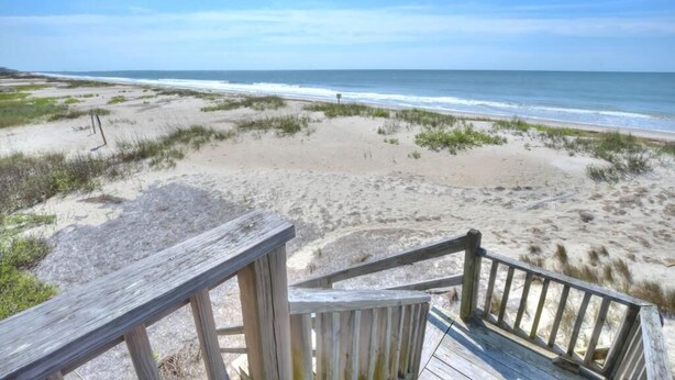 Une terrasse en bois qui mène à une plage bordée de dunes de sable et offrant une vue sur l’océan.