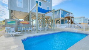 Una piscina con cubierta de madera y sillas de descanso, una estructura de madera con toldo azul, y una casa al fondo.
