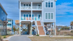 Una casa frente al mar de dos pisos, con un balcón en el nivel superior, un vestíbulo cubierto y una escalera que conduce a la entrada.