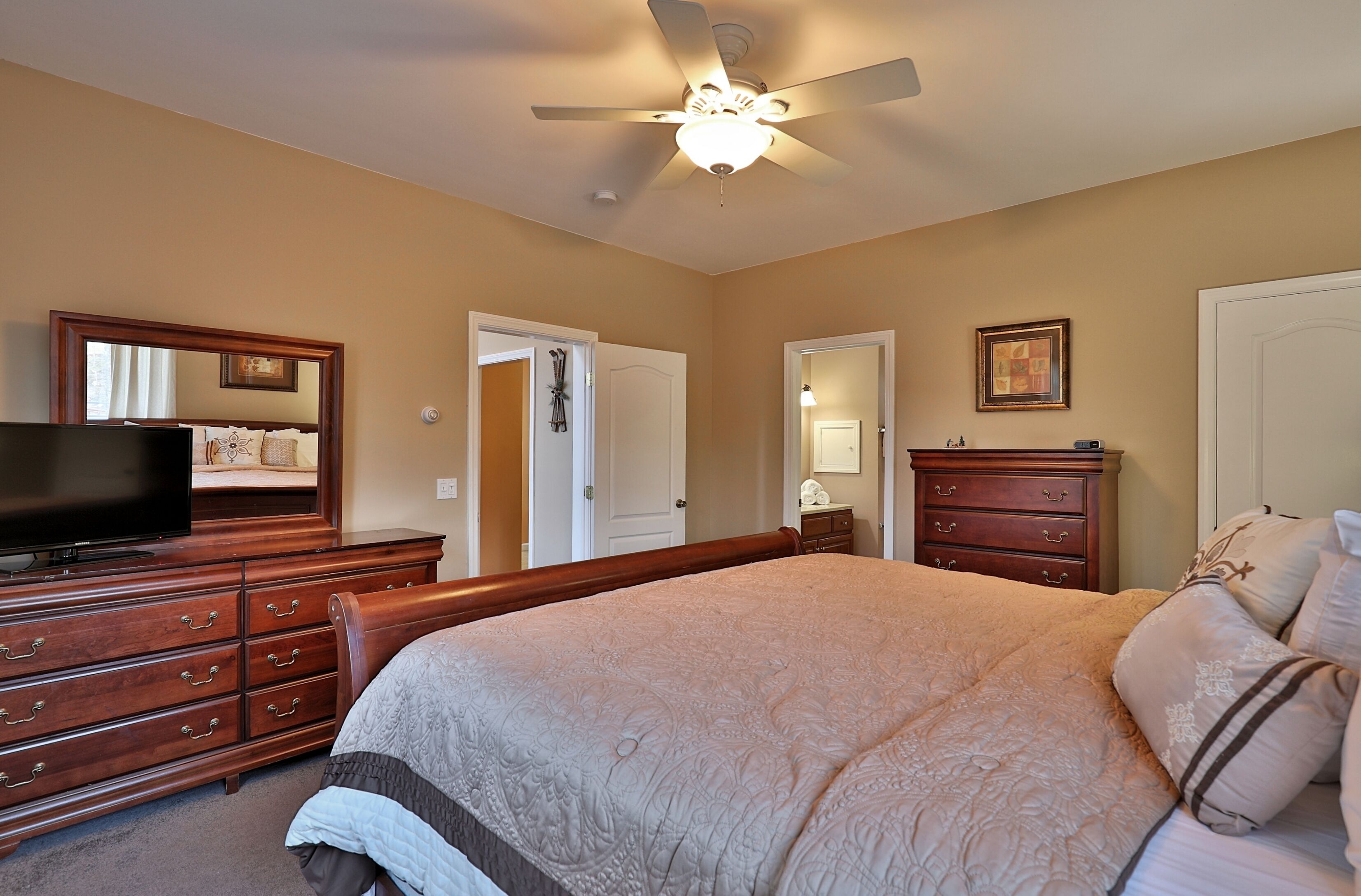 A bedroom with a large bed, wooden dresser, a television, and a ceiling fan.