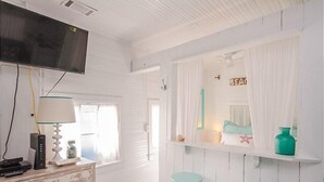 A room with a white wooden wall, a mounted TV, a desk with a lamp, two stools, and a shelf with decorative items. - Romantic 1930s Vintage Beach Cottage Steps to Beach by Tybee Cottages (Tybee Isand)