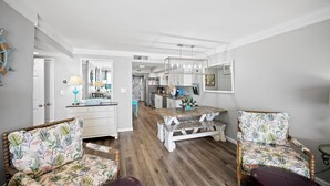 Amplia sala de estar con una mesa de comedor de madera, dos sillas con estampado floral, un banco y una cocina al fondo.