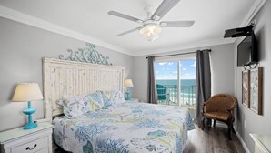 Un dormitorio con cama, ventilador de techo, una silla, televisión y vista al océano.