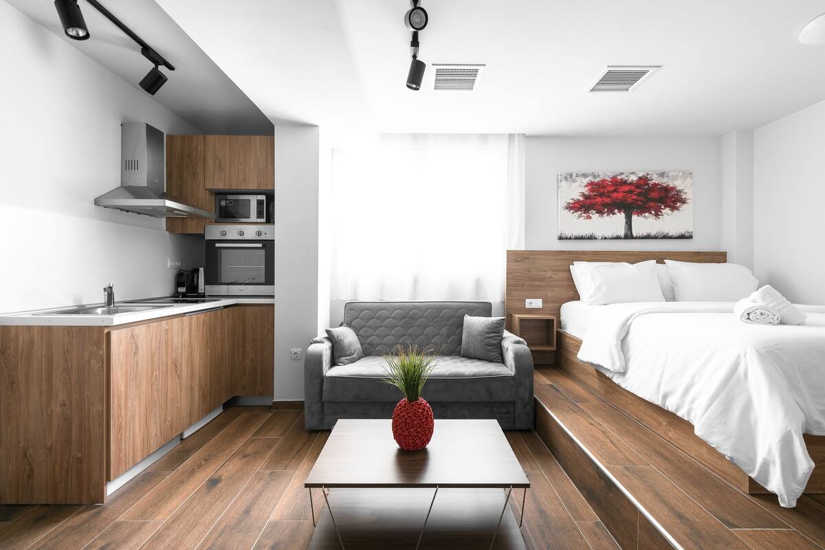 Una habitación de hotel moderna con kitchenette, un sofá gris, una cama con ropa blanca y un cuadro de un árbol.