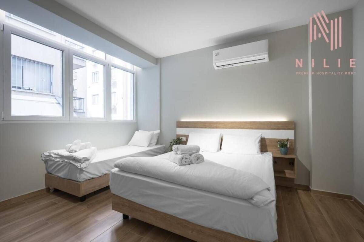 Habitación de hotel con dos camas, cabecero de madera, mesita de noche pequeña y unidad de aire acondicionado.