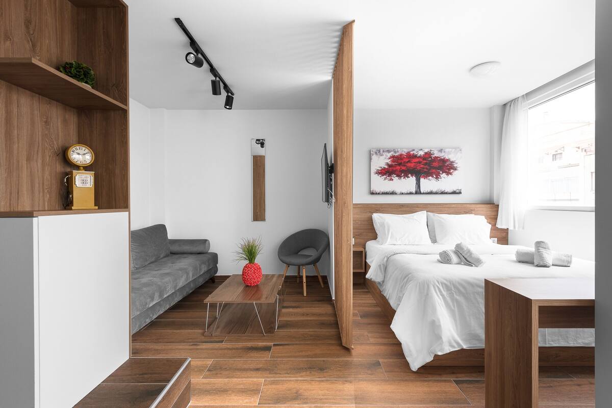 Una habitación moderna y funcional con piso de madera, un sofá gris, una mesita con una piña, una cama con ropa blanca y un cuadro de un árbol rojo.
