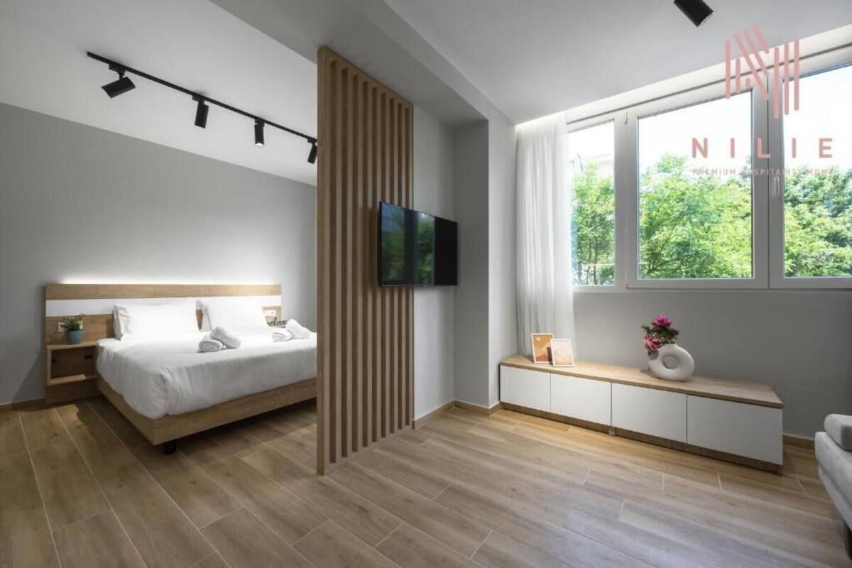 Un dormitorio moderno con cama, mesita de noche, televisor, ventana y vista a zonas verdes.