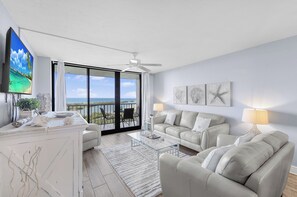 A modern living room with a balcony, white furniture, and artwork on the walls. - Beachfront Condo | Views of Beach, Pool & Grill (Marco Island)