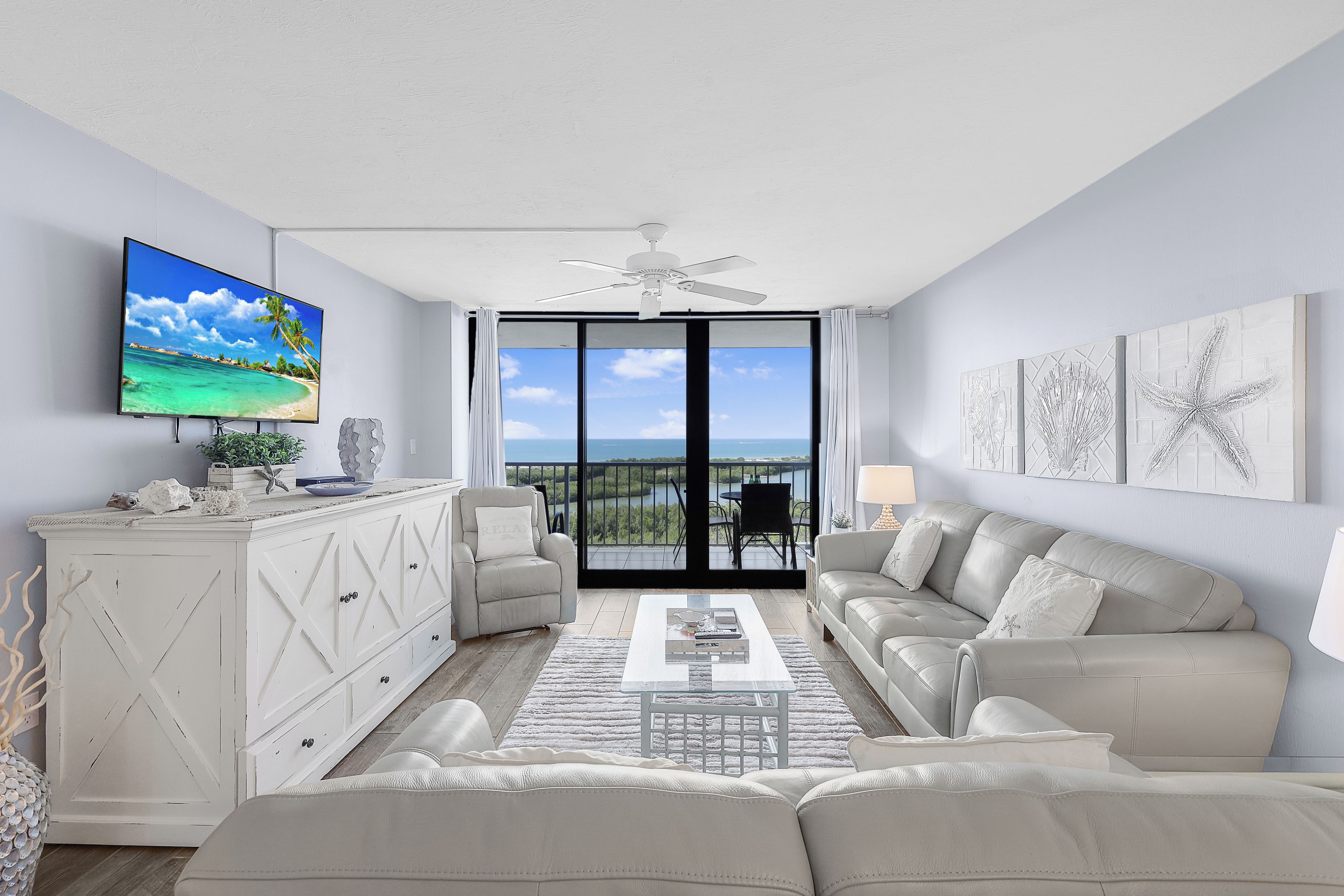 A modern living room with a large flat-screen TV, a ceiling fan, a white sofa, a coffee table, and a view of the ocean through large windows.