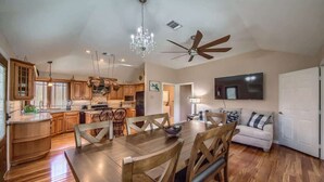 A spacious kitchen with wooden cabinets, a dining table, and chairs. There is a ceiling fan, a chandelier, and a television mounted on the wall. - PRD 2495 Ponderosa Country Haus (New Braunfels)