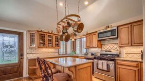 A kitchen with wooden cabinets, a central island, stainless steel appliances, and a hanging pot rack. - PRD 2495 Ponderosa Country Haus (New Braunfels)