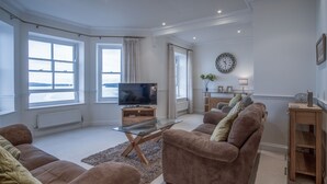 Ein geräumiges Wohnzimmer mit großem Flachbildfernseher, bequemen Sofas, einem Glastisch und großen Fenstern mit Blick.
