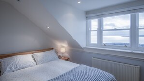 Ein Schlafzimmer mit Bett, Nachttisch, Lampe, Fenster mit Blick und Heizkörper.