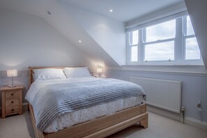 A bedroom with a sloped ceiling, a large window, a wooden bed, a nightstand, and a wall-mounted radiator. - South Beach View- 2 Bedroom - Tenby (Tenby)