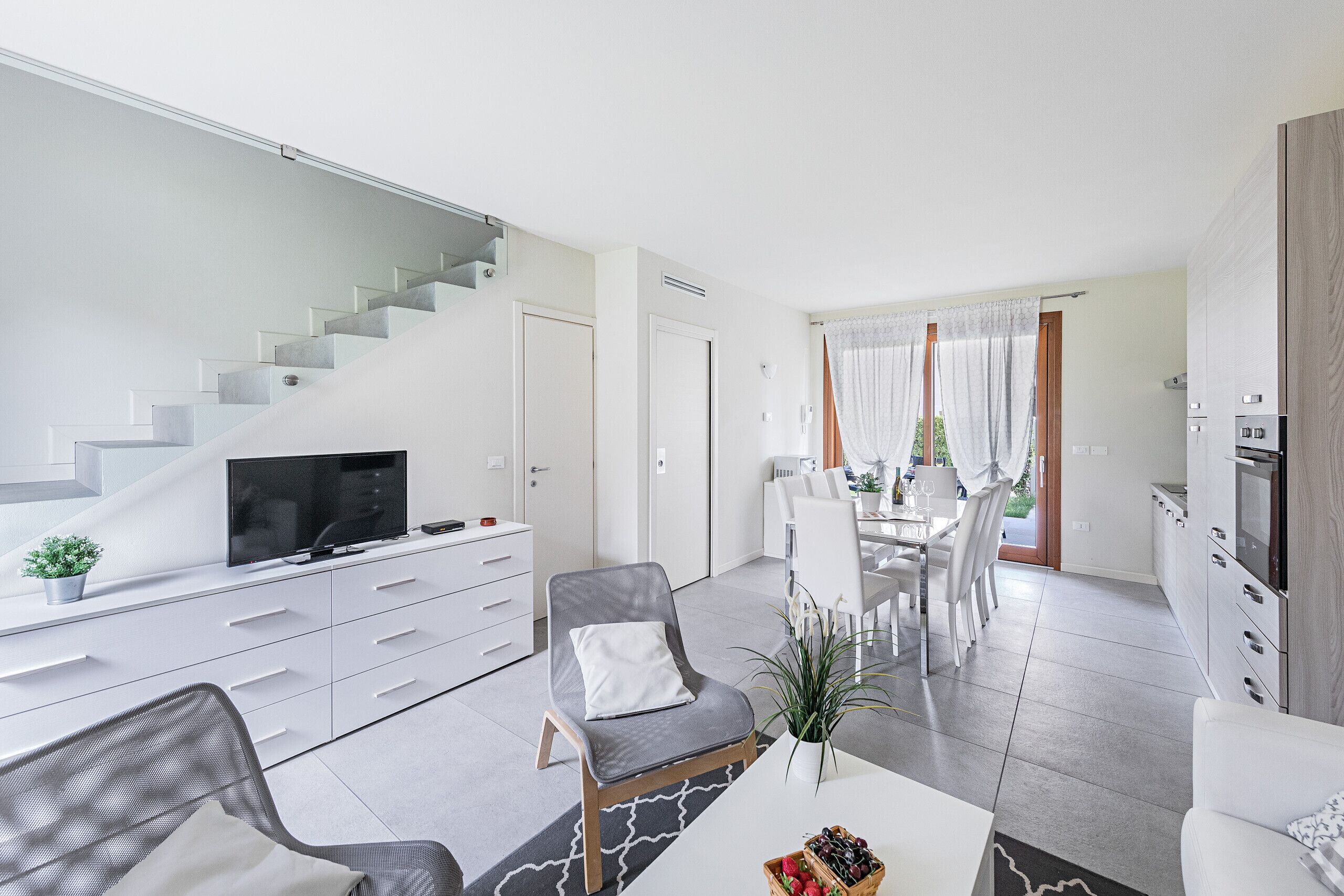 A modern living room with a flat-screen TV, a dining area with white furniture, and a kitchen with built-in appliances.