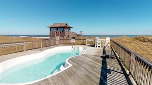 Una terraza de madera con piscina, una torre de salvavidas y sillas de playa.