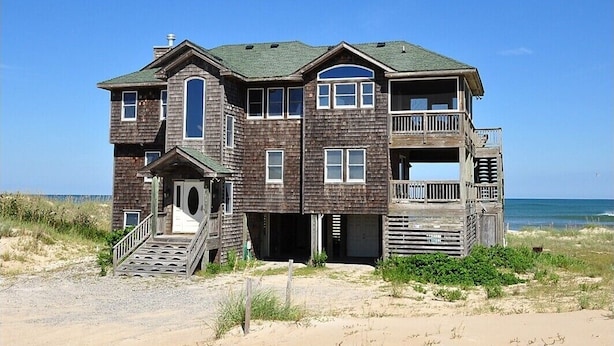 Una casa de playa de dos pisos, con un balcón, un porche delantero y una escalera que conduce a la entrada.
