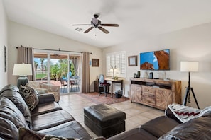 A living room with a sofa, ottoman, desk, television, and a ceiling fan.