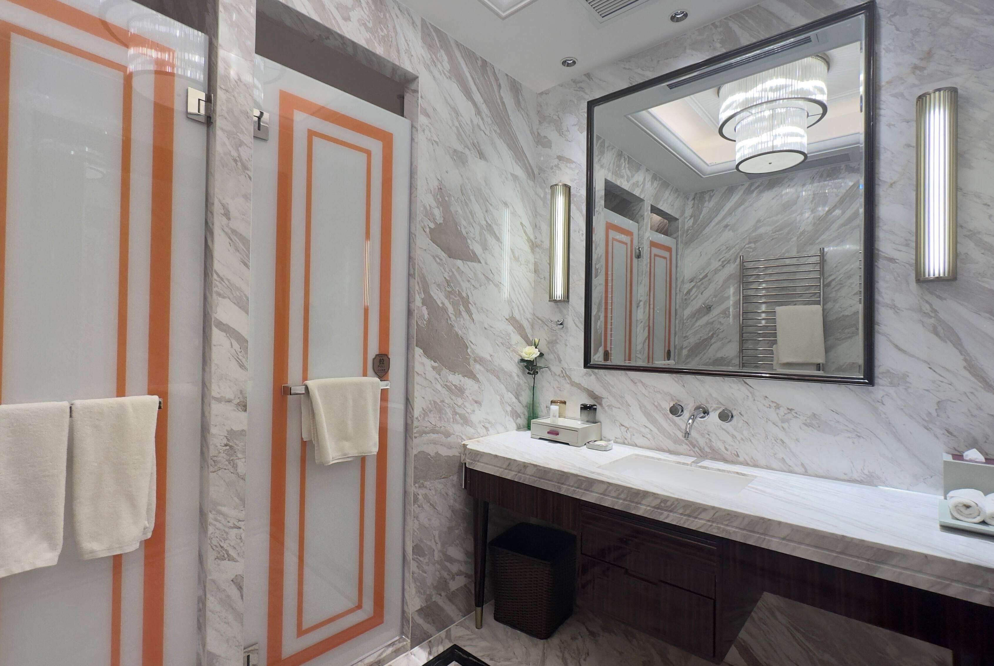 Une salle de bain aux murs recouverts de marbre, avec un grand miroir, un lavabo et deux serviettes accrochées au mur.