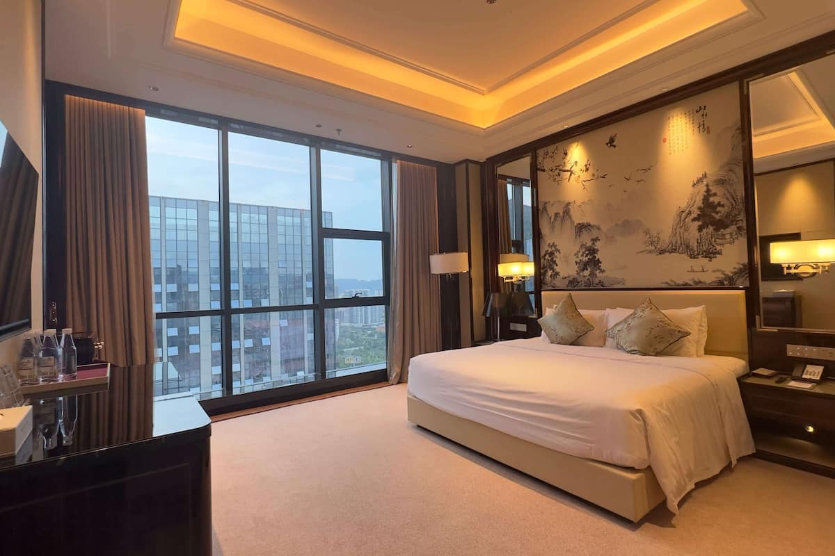 Ein Hotelzimmer mit einem großen Bett, einem Schreibtisch mit Stuhl, einem großen Fenster mit Vorhängen und einer Wand mit einem traditionellen Gemälde.