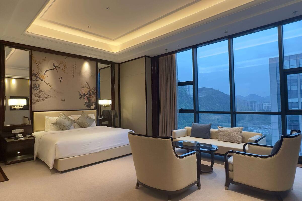 Ein Hotelzimmer mit einem großen Bett, einem Schlafsofa, einem Couchtisch und Blick auf die Stadt durch große Fenster.