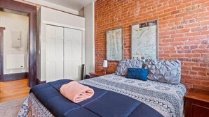 A bedroom with a brick wall, a bed with patterned bedding, a folded towel, and two maps on the wall. - Chic 2BR: Near Downtown, Copper Mountain (Leadville)