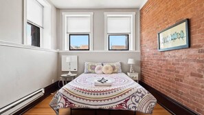 A bedroom with a large bed, two windows, a brick wall, and a framed artwork. - Chic 2BR: Near Downtown, Copper Mountain (Leadville)