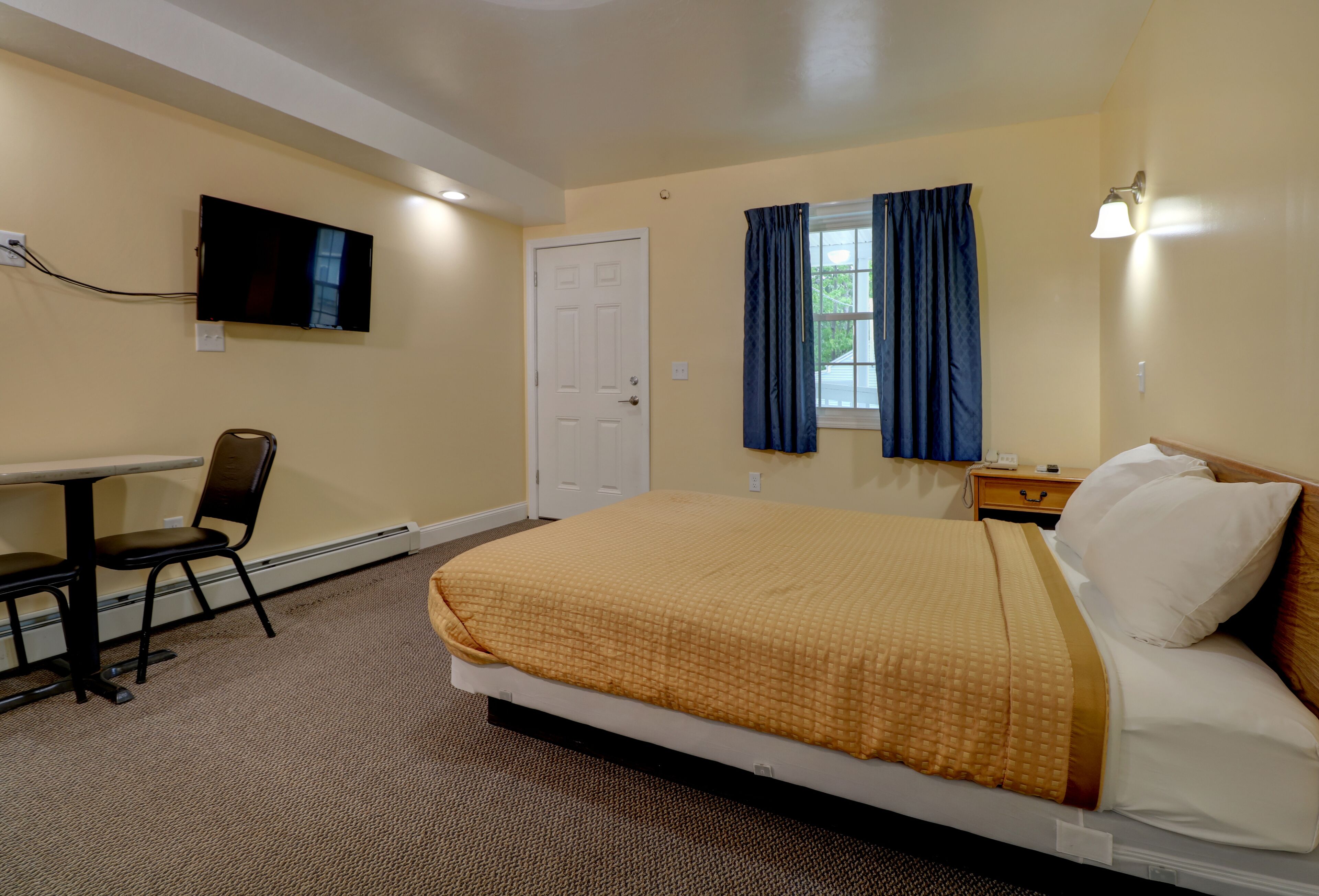 Habitación de hotel con cama, escritorio, silla, televisor, ventana con cortinas y mesita de noche.