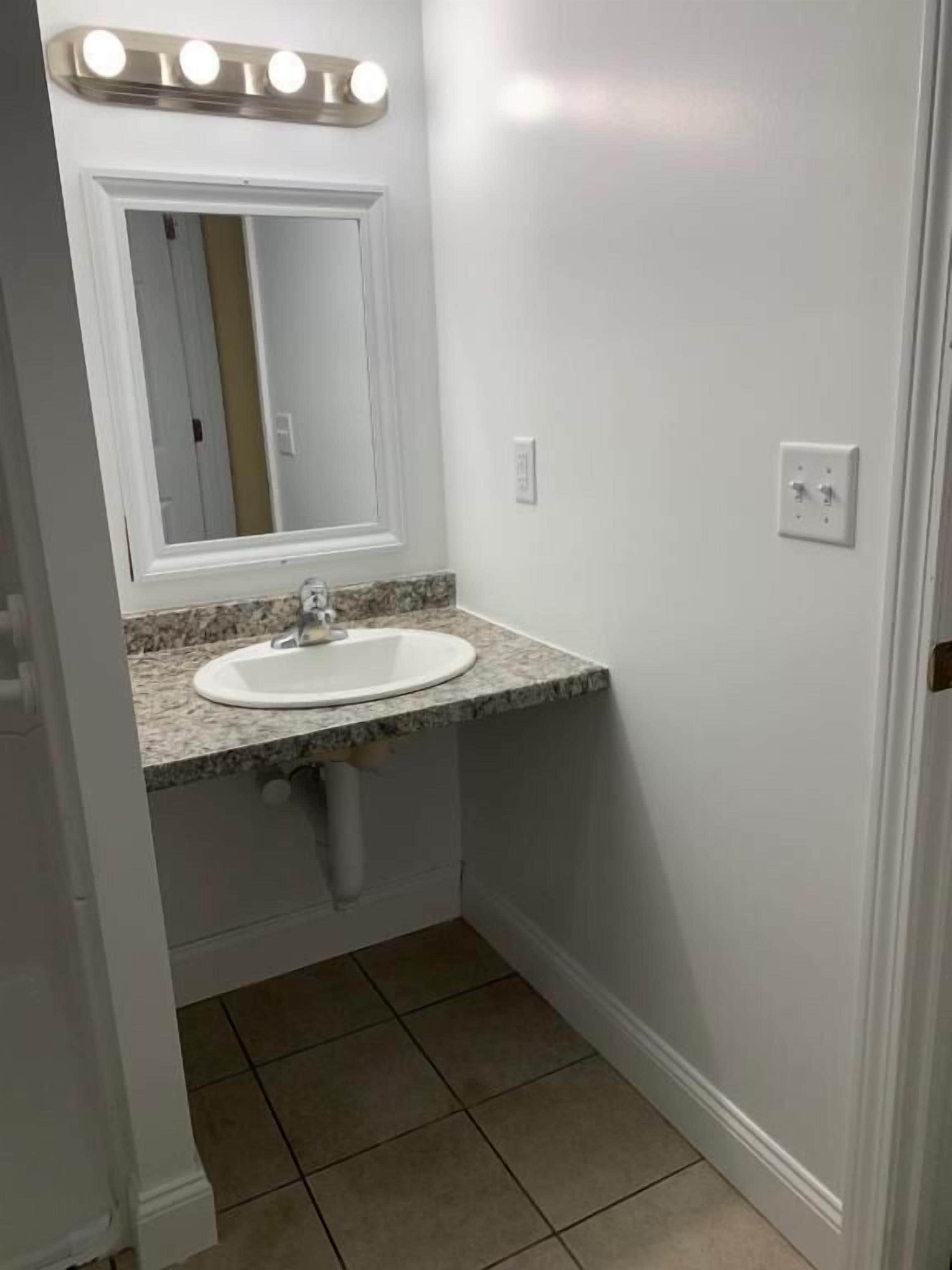 A bathroom with a white sink, granite countertop, and a mirror with lights above.