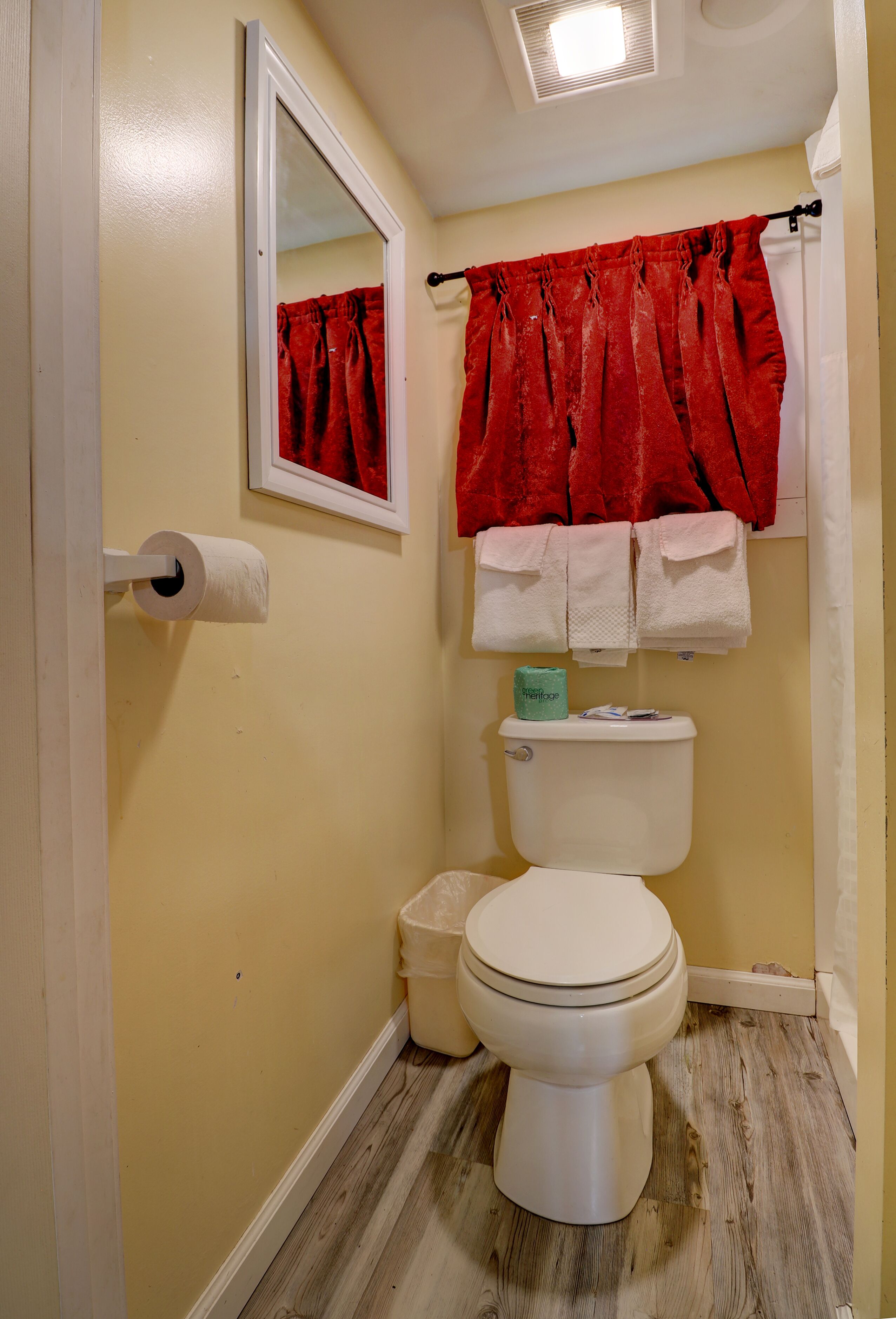 A small bathroom with a toilet, a roll of toilet paper, a trash bin, and a red shower curtain.