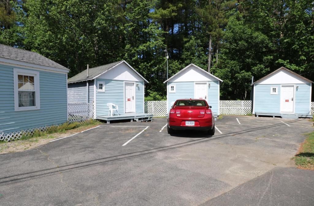 A row of light blue cabins with white trim, each featuring a small porch and a door. A red car is parked in the middle of the parking lot.
