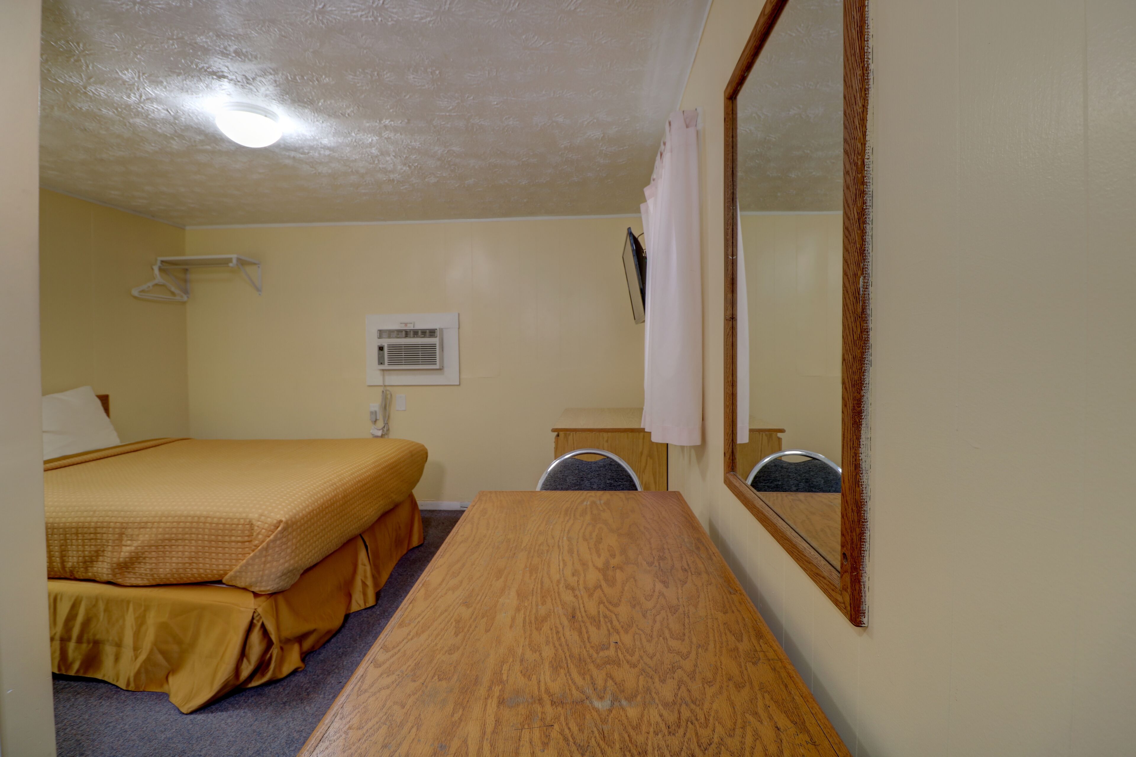 A hotel room with a wooden desk, a bed with yellow bedding, a mirror, and a wall-mounted air conditioning unit.