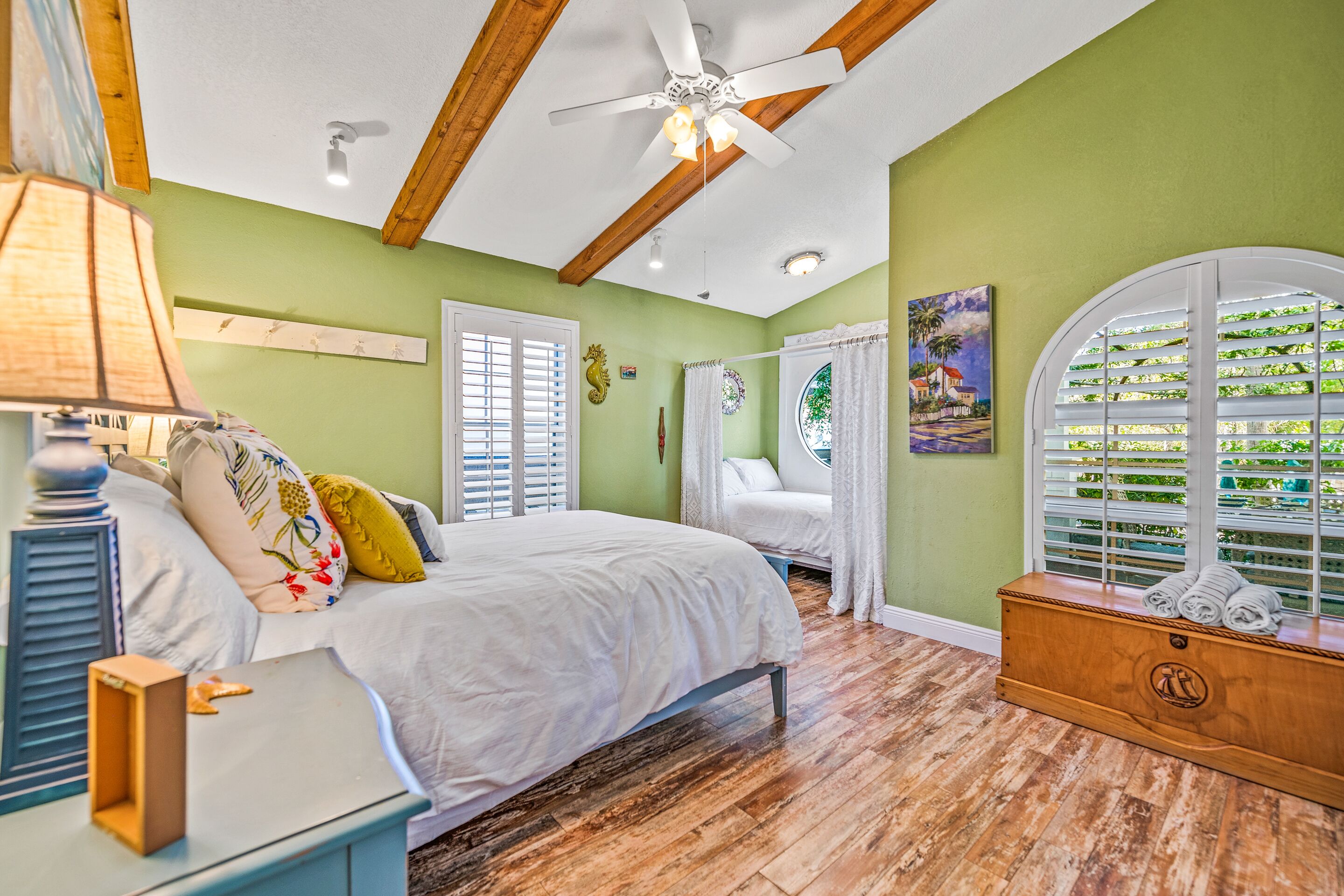 A bedroom with a bed, a nightstand, a wooden dresser, a ceiling fan, and a window with shutters.