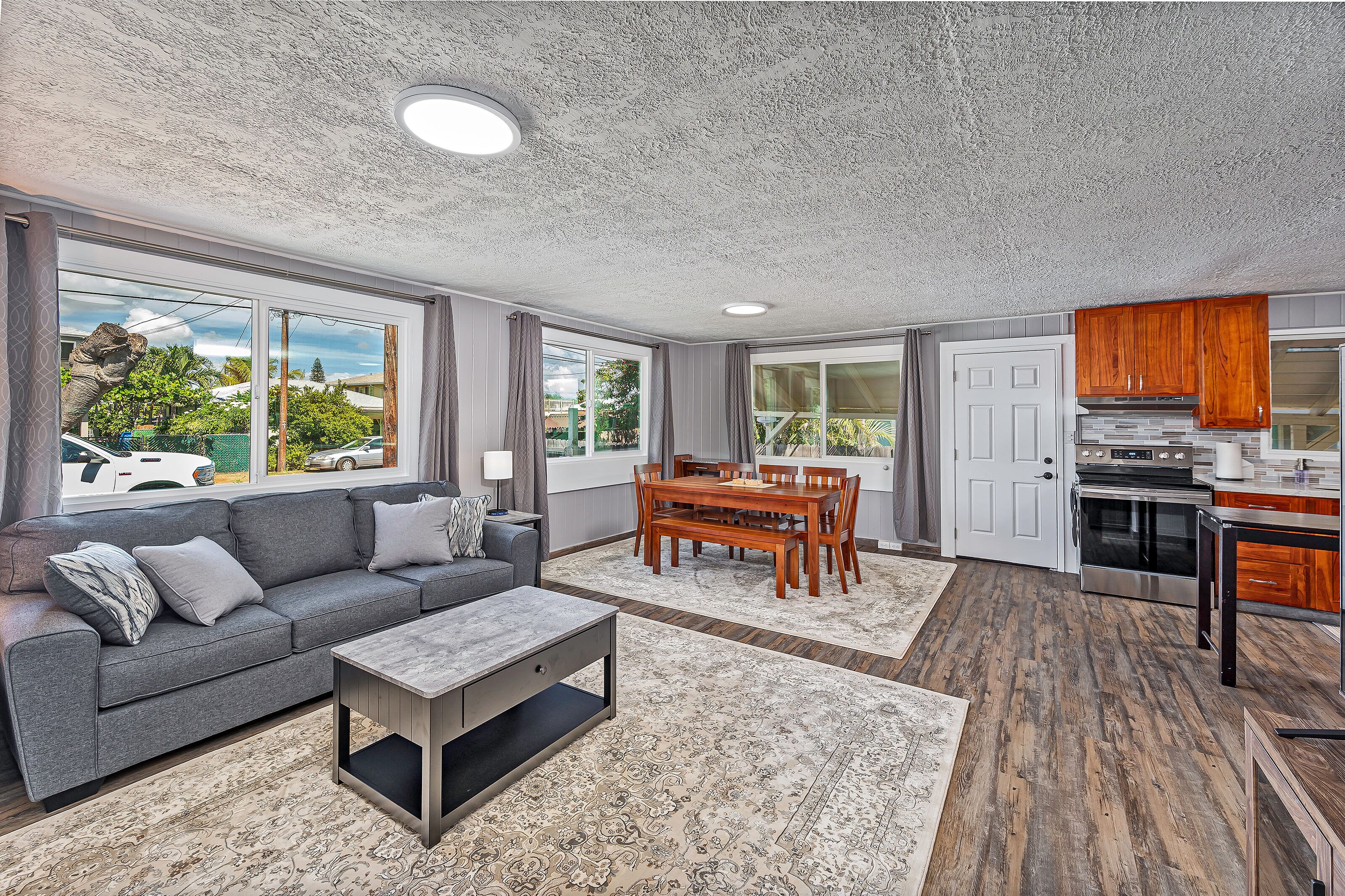 A modern living room with a grey sofa, wooden dining table, and a kitchen area with wooden cabinets and appliances.