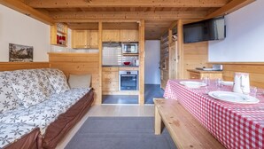 A cozy cabin with a wooden interior, a bed with a floral bedspread, a dining table set for two, and a kitchenette with a microwave and oven. - Bel Apt Near The Slopes Argentiere (Chamonix-Mont-Blanc)