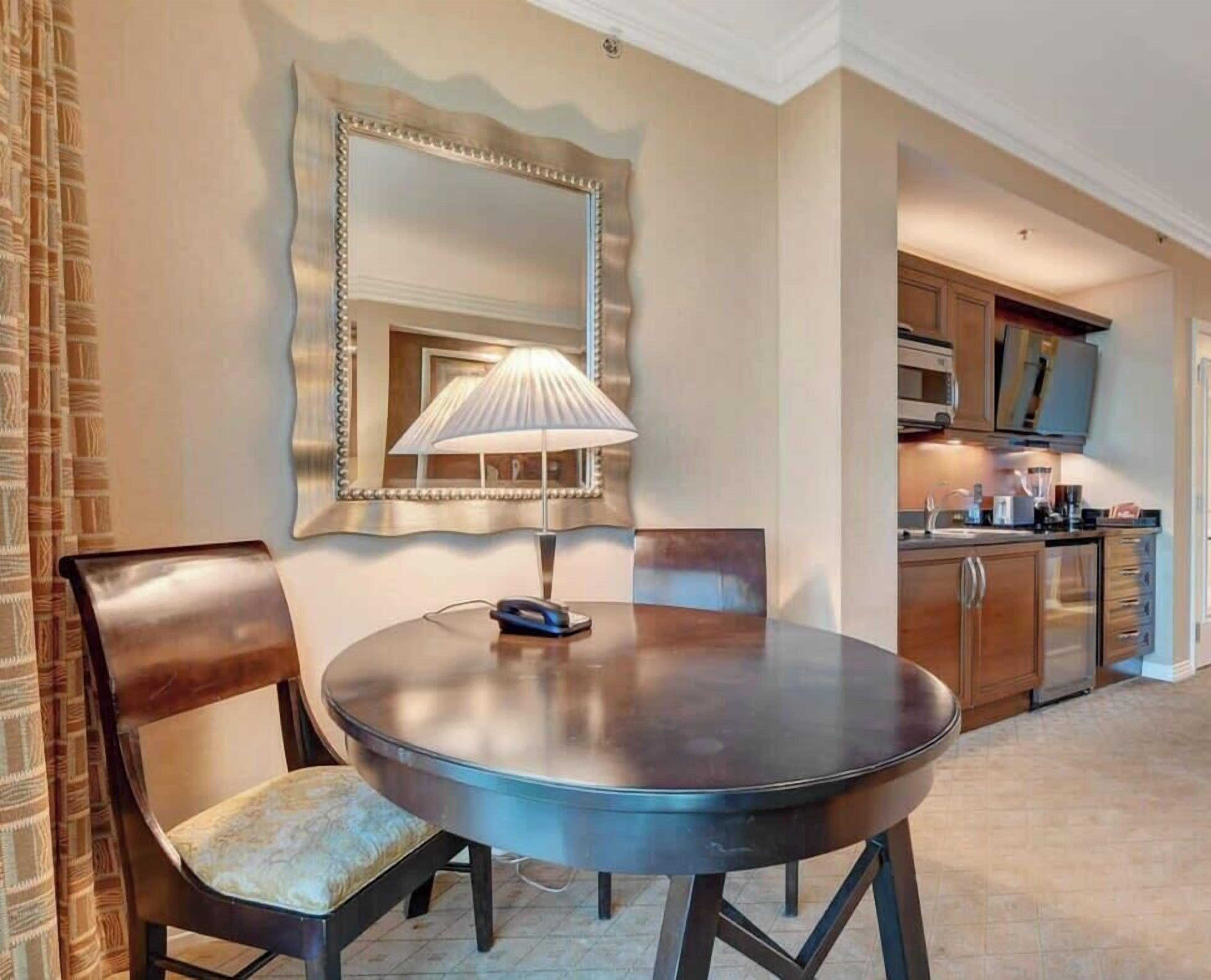 A dining area with a round table, two chairs, a lamp, and a kitchen in the background.