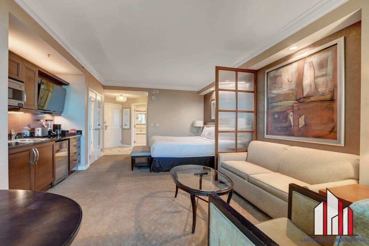 A hotel room with a bed, a sofa, a dining table, a kitchen area, and a large painting on the wall.