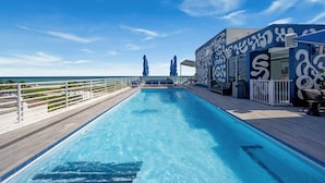 Ein Poolbereich mit freiem Blick auf den Ozean, eine Terrasse mit Sitzgelegenheiten und ein Gebäude mit markanter Außenarchitektur.