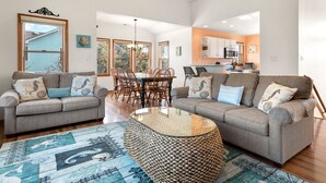 A spacious living room with two grey sofas, a glass coffee table, and a dining area with wooden chairs. - Ducktopia: 6-minute walk to Beach, Pool, Hot Tub, Short Walk to Downtown Duck (Duck)