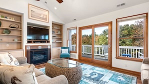 A living room with a fireplace, television, and a view of the outdoors through large windows. - Ducktopia: 6-minute walk to Beach, Pool, Hot Tub, Short Walk to Downtown Duck (Duck)