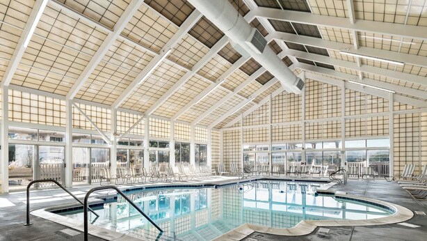 An indoor swimming pool with clear glass walls, a high ceiling with a grid pattern, and multiple lounge chairs around the pool area. - Ducktopia: 6-minute walk to Beach, Pool, Hot Tub, Short Walk to Downtown Duck (Duck)