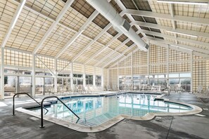 An indoor swimming pool with clear glass walls, a high ceiling with a grid pattern, and multiple lounge chairs around the pool area. - Ducktopia: 6-minute walk to Beach, Pool, Hot Tub, Short Walk to Downtown Duck (Duck)