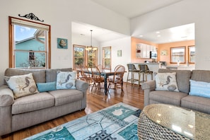 A spacious living room with two sofas, a dining table, and chairs. A kitchen is visible in the background with modern appliances.