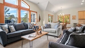 A spacious living room with a grey sofa, a white sofa, and a grey armchair. There is a wooden coffee table with a lamp and candles. Large windows provide natural light and a view of the outdoors. - Ocean Escape: Lakefront, Pool, Hot Tub, Game Room (Corolla)