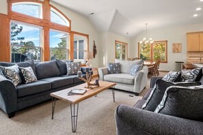 Un salon spacieux avec un canapé gris, un canapé blanc et un fauteuil gris. On y trouve une table basse en bois, sur laquelle sont posés une lampe et des bougies. De grandes fenêtres laissent entrer la lumière naturelle et offrent une vue sur l’extérieur.